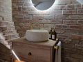 A modern bathroom featuring Covent beige floor tiles, light Casella oak vanity units, brushed brass fittings, freestanding oval bath, and round LED backlit mirror.