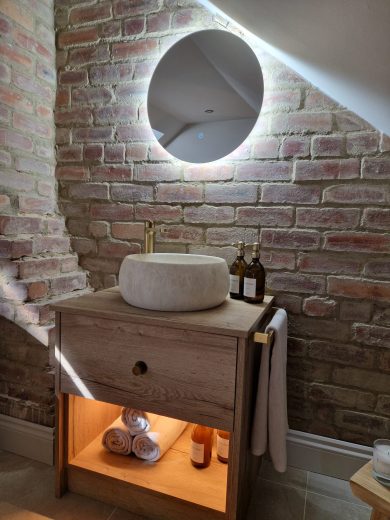 A modern bathroom featuring Covent beige floor tiles, light Casella oak vanity units, brushed brass fittings, freestanding oval bath, and round LED backlit mirror.