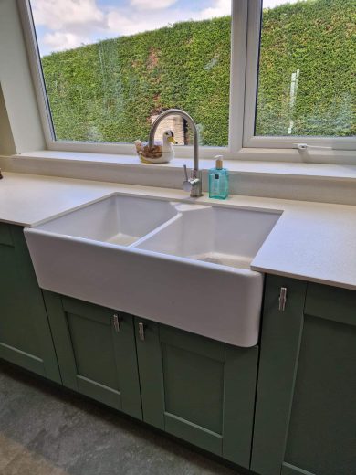 A bespoke kitchen with green shaker-style cabinets, white worktops, Belfast sink, and modern appliances designed by Dragonville Interiors.