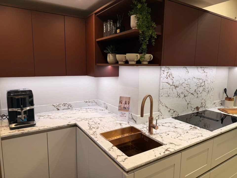 Modern fitted kitchen with marble-effect worktops, copper sink and tap, induction hob, and matte brown wall cabinets with open shelving and under-cabinet lighting.