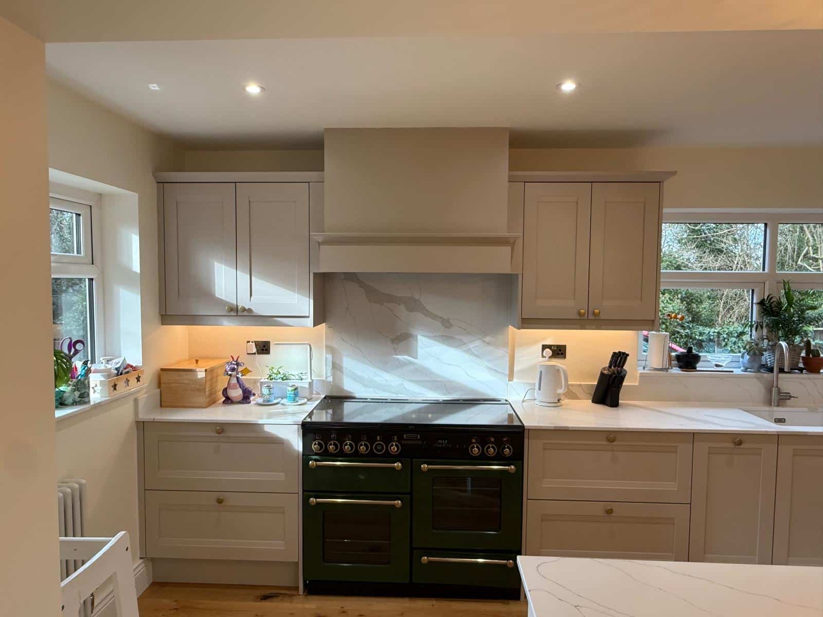A bespoke kitchen in warm neutral tones with shaker-style cabinetry, marble-effect worktops, range cooker, and soft under-cabinet lighting by Dragonville Interiors.